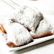A plate of golden-brown New Orleans beignets generously dusted with powdered sugar sits on a white surface. The pastries look soft, square-shaped, and freshly made.