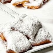 A plate of square New Orleans beignets generously dusted with powdered sugar, with more beignets in the background on a white surface.