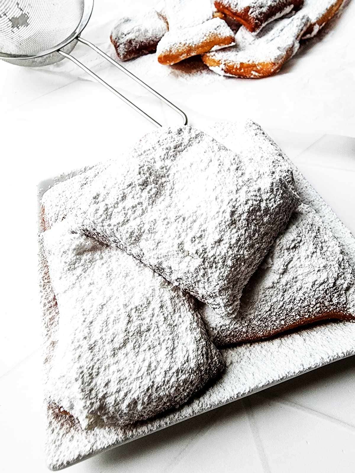 A plate of square New Orleans beignets generously dusted with powdered sugar, with more beignets and a fine mesh strainer in the background on a white surface.