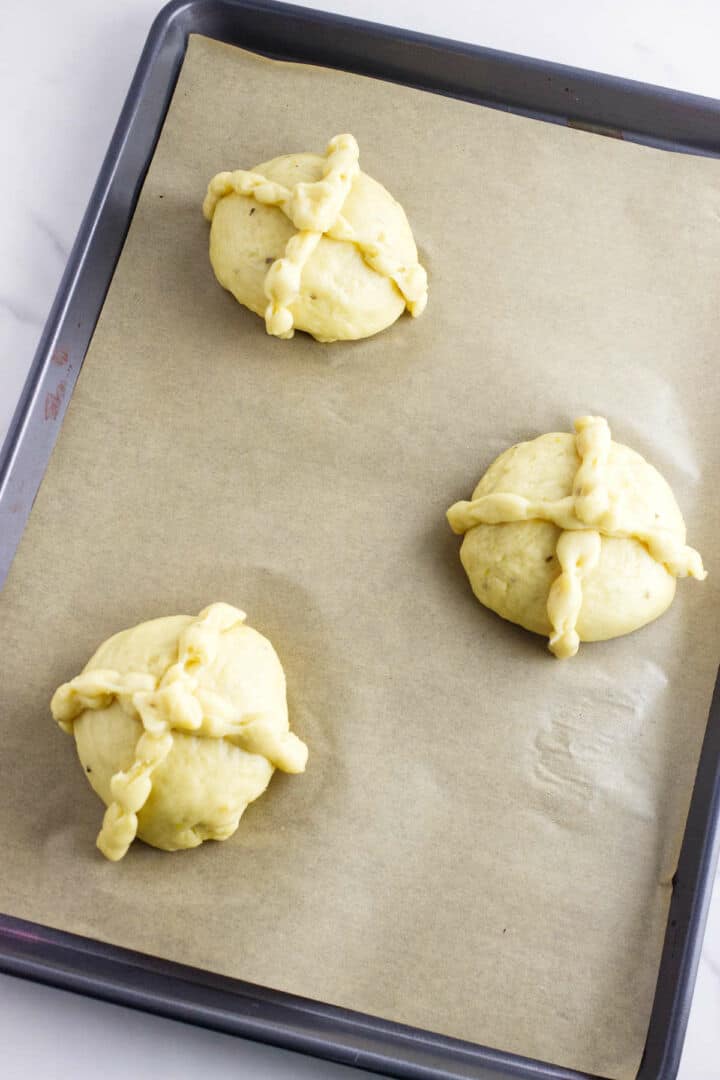 Three unbaked round dough balls with braided cross patterns on top are placed on a parchment-lined baking sheet, ready to be baked as traditional pan de muerto for Day of the Dead celebrations.