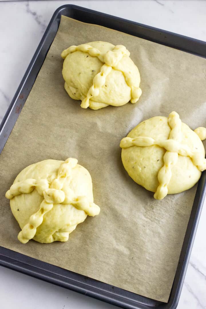 Three round pan de muerto dough balls with crisscrossed strips on top sit on a parchment-lined baking tray, ready to be baked. The light yellow dough is smooth and perfectly shaped for this traditional Day of the Dead bread.