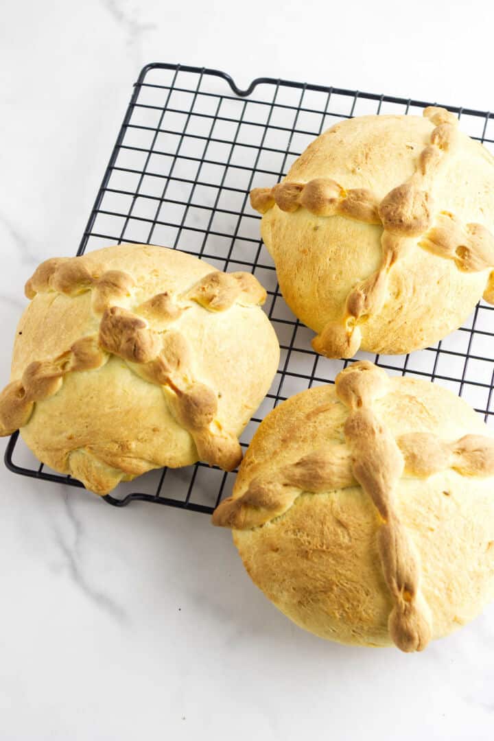 Three round loaves of pan de muerto, traditional Day of the Dead bread with braided crosses on top, are cooling on a black wire rack on a white marble surface.