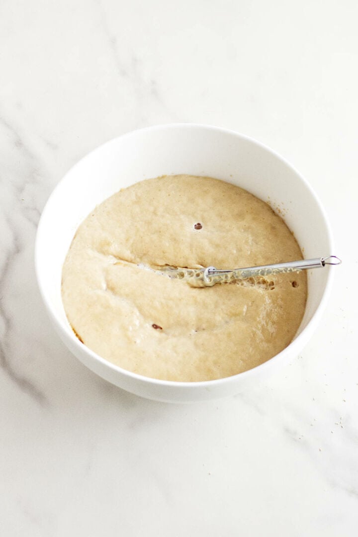 A white bowl filled with bubbly, risen pan de muerto dough sits on a white marble surface. A metal spoon rests inside the bowl, partially submerged in the traditional Day of the Dead bread mixture.