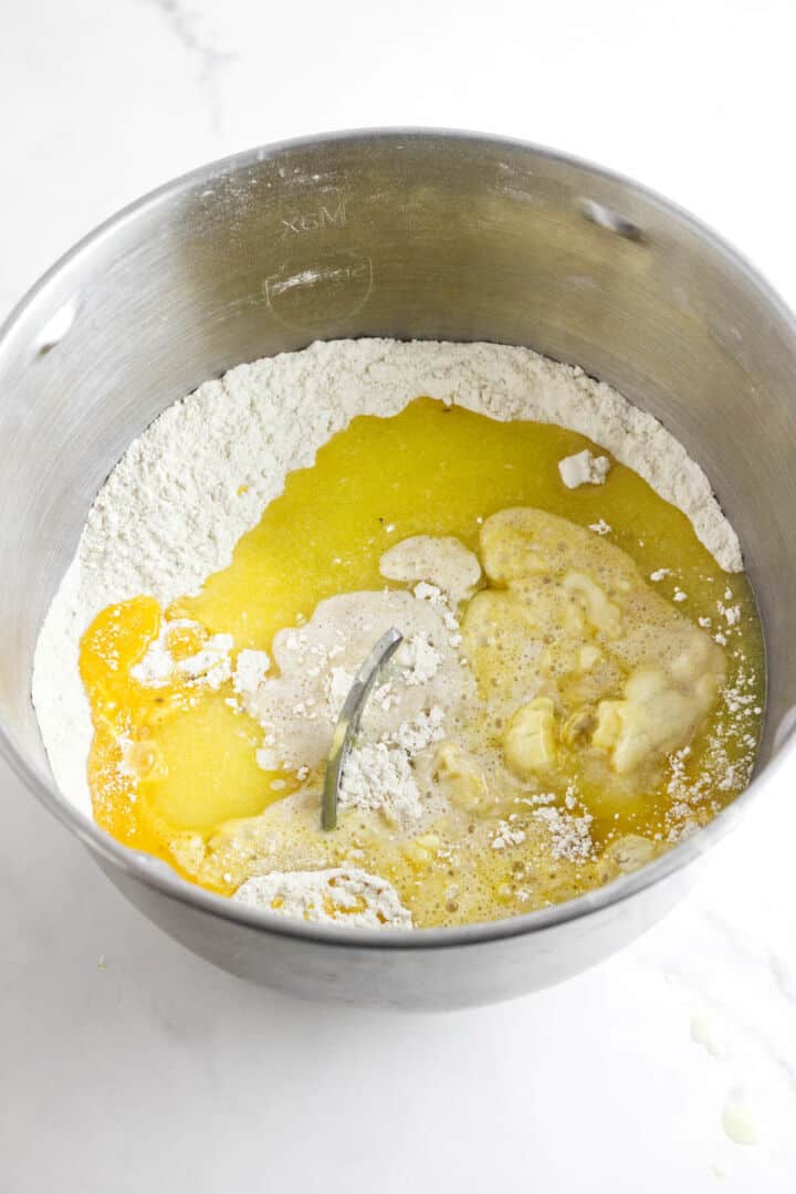 A metal mixing bowl contains flour, melted butter, eggs, and other wet ingredients to make day of the dead bread. The pan de muerto ingredients are ready for mixing but have not yet been combined.