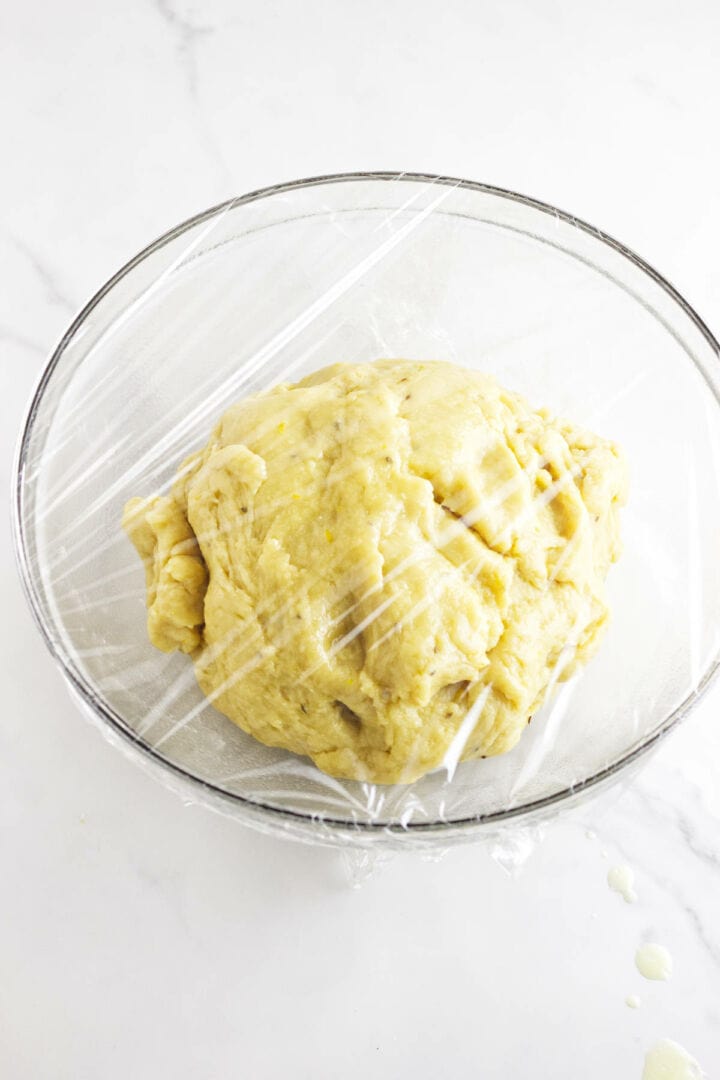 A glass bowl filled with pan de muerto dough, covered with plastic wrap, sits on a white marble surface.