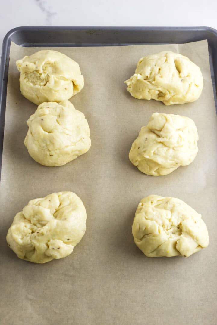 Six uneven portions of raw pan de muerto dough are placed on a baking sheet lined with parchment paper, ready to be baked. The dough is light yellow and has a rough texture.