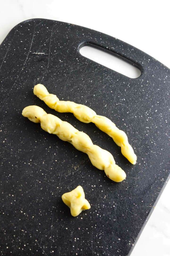 Two twisted strips and one small, knotted piece of pan de muerto rest on a black, speckled cutting board with a handle, on a white surface.