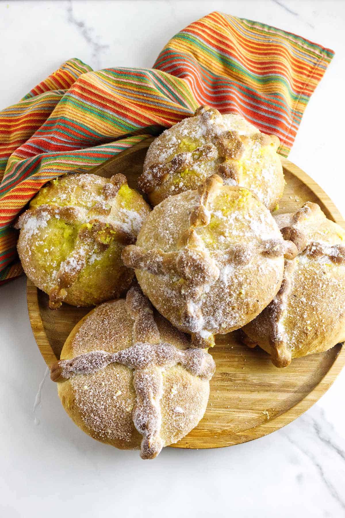 A wooden plate holds five round loaves of pan de muerto, traditional Day of the Dead bread dusted with sugar, next to a colorful striped cloth on a white marble surface.