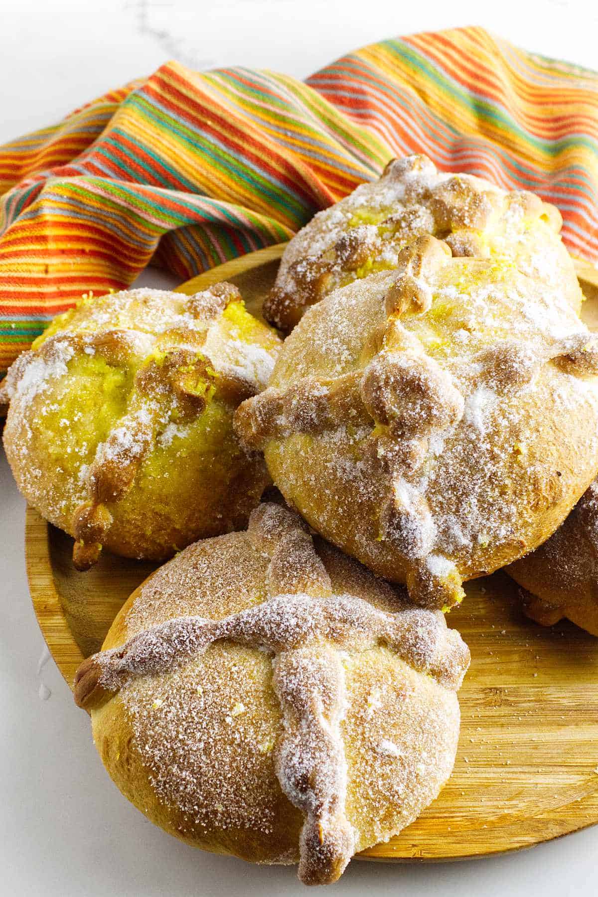 A wooden plate with several pieces of pan de muerto, the traditional Day of the Dead bread topped with sugar, is displayed in front of a colorful, striped cloth.
