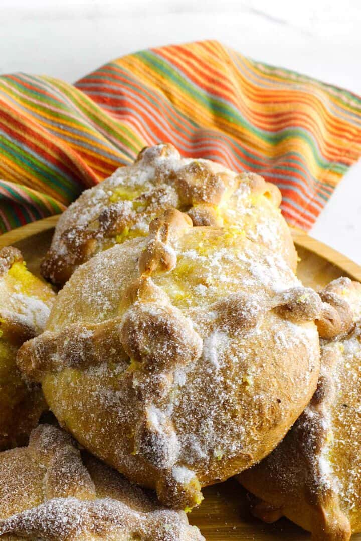 A close-up of pan de muerto, traditional Day of the Dead bread, topped with bone-shaped dough pieces and dusted with sugar, sits on a wooden plate with a colorful striped cloth in the background.