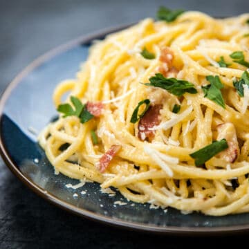 A close-up of spaghetti carbonara, one of the most beloved pasta recipes, topped with grated cheese, parsley, and pieces of bacon on a dark, round plate.