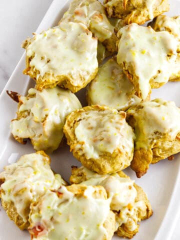 A white rectangular plate with a pile of round, golden persimmon cookies topped with a thick, glossy, white glaze. The cookies have an uneven, rustic shape and are arranged closely together.