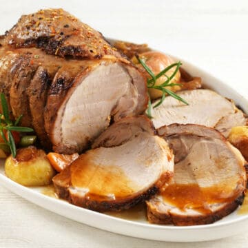 A roasted pork loin is served on a platter with slices cut, surrounded by roasted potatoes, carrots, onions, and sprigs of fresh rosemary, topped with a glossy brown gravy—perfect for fans of classic pork recipes.