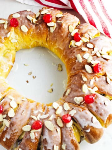 A golden-brown rosca de reyes Christmas cake, ring-shaped and topped with white icing, red cherries, and sliced almonds, sits on a white surface near a red-and-white striped towel.