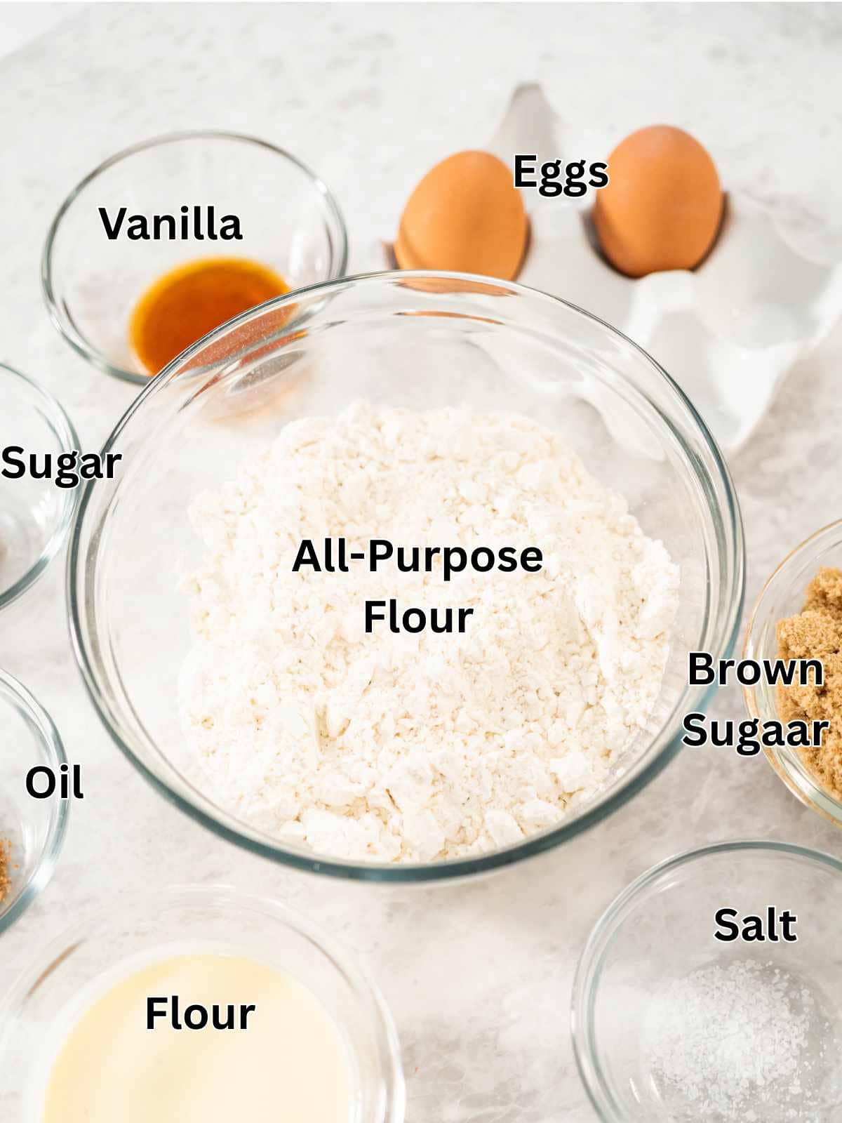 A glass bowl labeled "All-Purpose Flour" surrounded by small bowls of vanilla, eggs, sugar, brown sugar, oil, flour, and salt on a white surface-everything you need to start baking a classic Texas tornado cake.