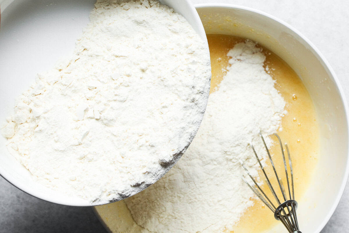 A bowl of flour is being poured into a larger bowl containing a yellow batter for a Texas tornado cake, with a metal whisk resting inside the larger bowl.