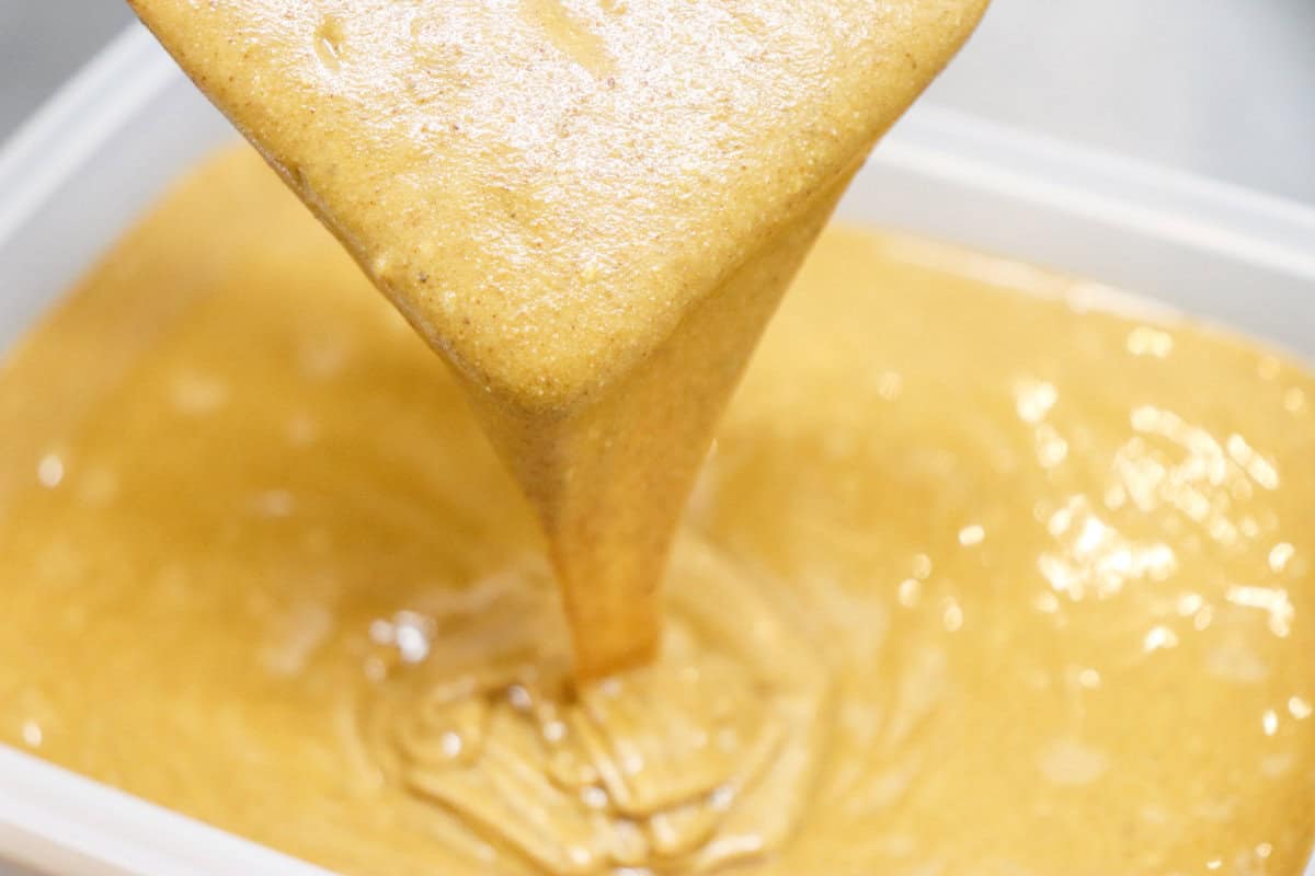 Thick, golden-brown batter is being poured into a container, forming swirls on the surface. The smooth, creamy texture hints that it might be for baking a decadent Texas Tornado Cake.