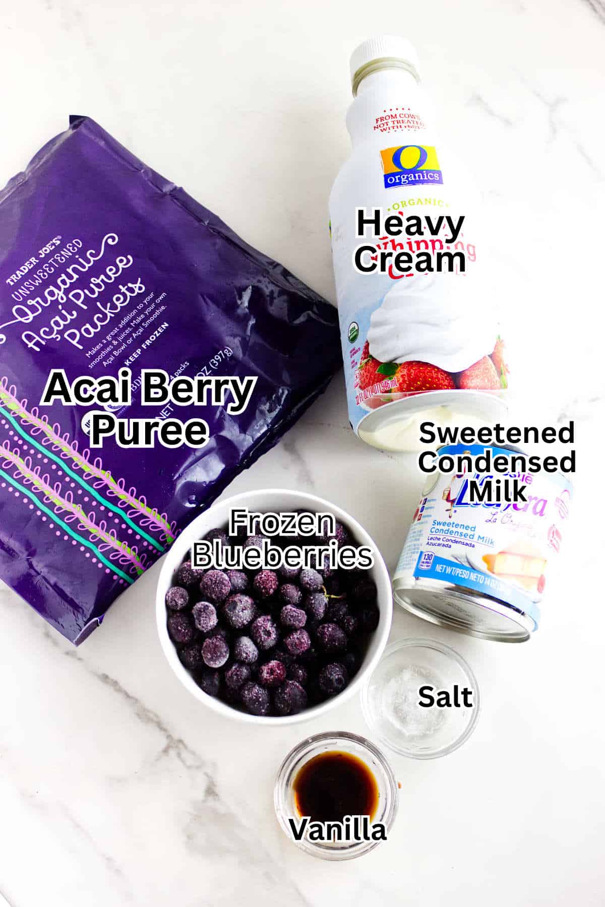 Acai berry puree packet, bottle of heavy cream, can of sweetened condensed milk, and bowl of frozen blueberries—all the essentials for making delicious acai berry ice cream—are labeled and arranged on a white surface.