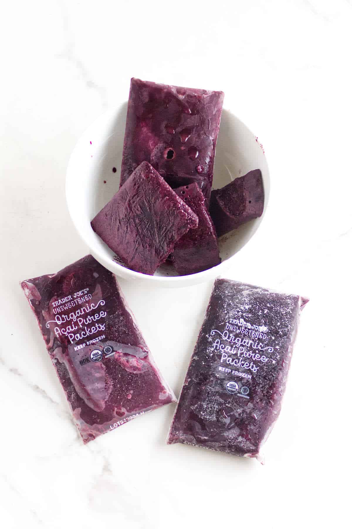 A white bowl filled with dark purple frozen acai packets sits on a white surface, resembling acai berry ice cream, with two unopened Trader Joe's organic acai puree packets lying in front of the bowl.