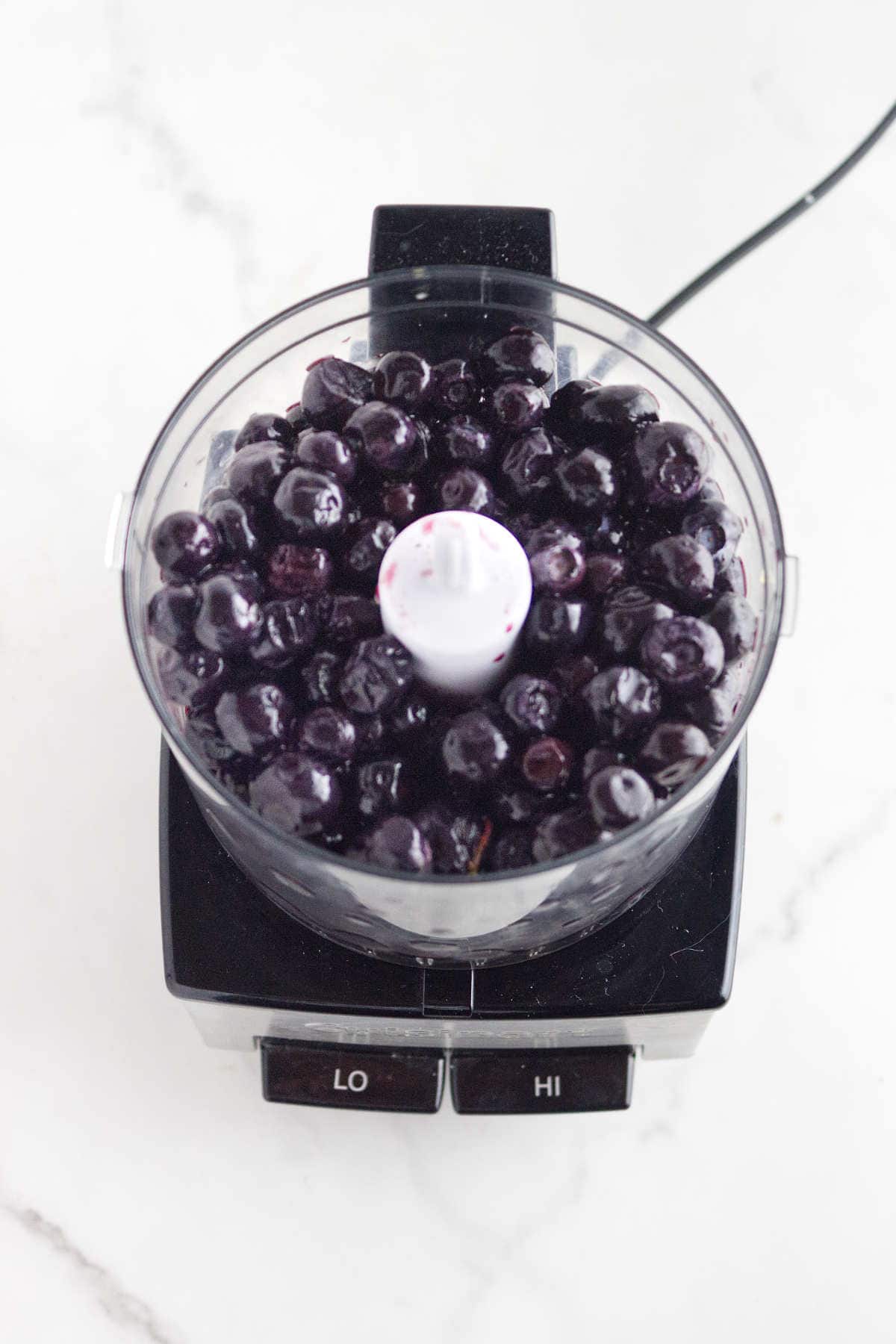 A food processor filled with fresh blueberries sits on a white countertop, viewed from above-perfect for whipping up homemade acai berry ice cream using the "LO" and "HI" settings.
