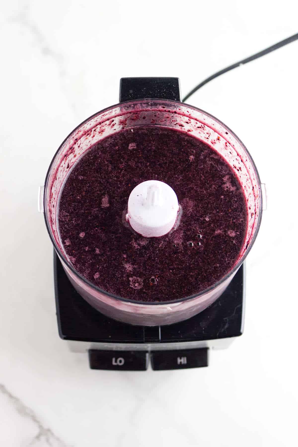 A food processor filled with a dark purple acai berry ice cream mixture sits on a white countertop, viewed from above. The base has two buttons labeled "LO" and "HI.