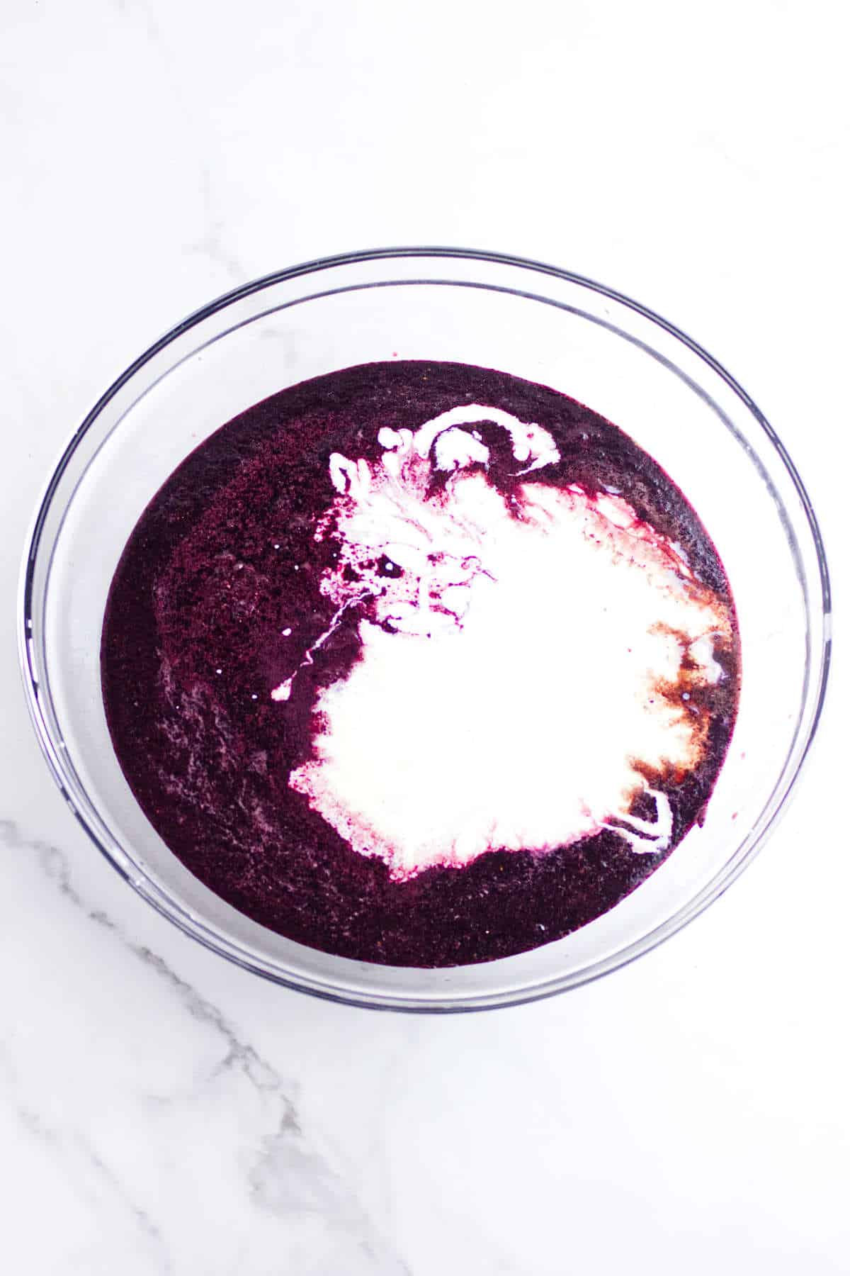 A glass bowl filled with a deep purple acai berry ice cream mixture and a swirl of white liquid on top, placed on a white marble surface.