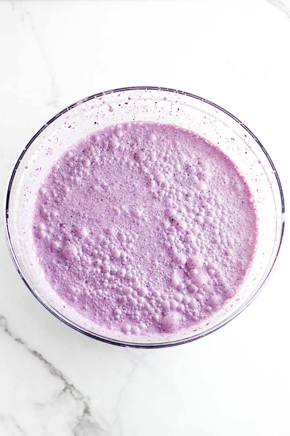 A clear glass bowl filled with a frothy, bubbly purple liquid, reminiscent of acai berry ice cream, sits on a white marble surface.