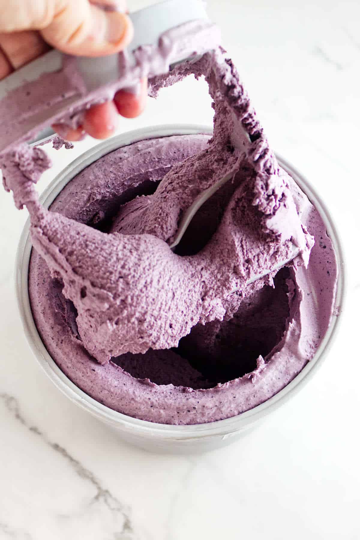 A hand holds a mixer attachment covered in thick, creamy acai berry ice cream above a bowl filled with the same vibrant purple mixture on a white marble surface.