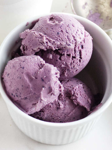 A white bowl filled with three scoops of acai berry ice cream sits on a white surface, next to a metal ice cream scoop.