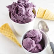 Two cups filled with purple acai berry ice cream, one large and one small, with a metal ice cream scoop and two empty waffle cones on a white surface.