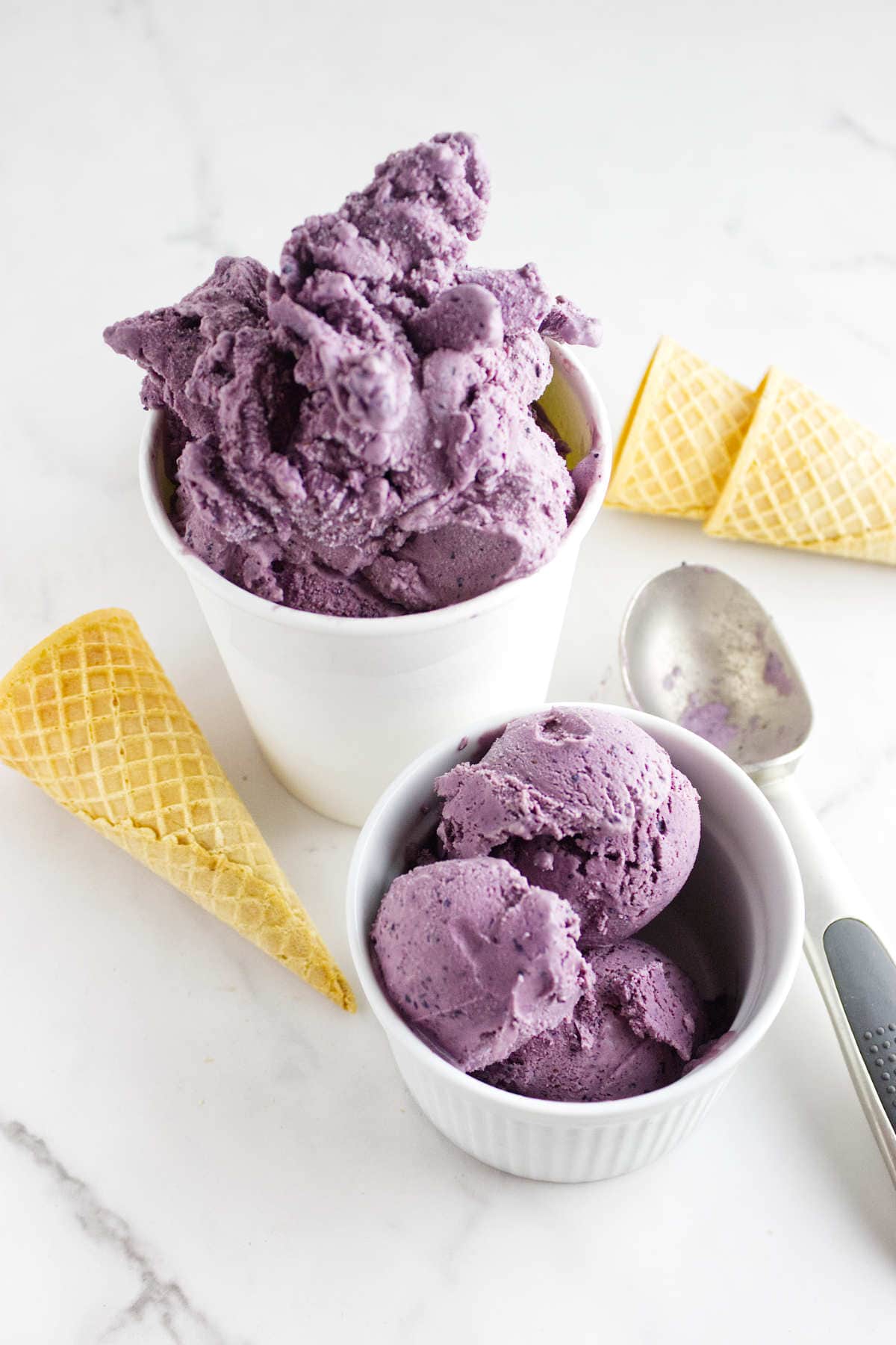 Two cups filled with purple acai berry ice cream, one large and one small, with a metal ice cream scoop and two empty waffle cones on a white surface.