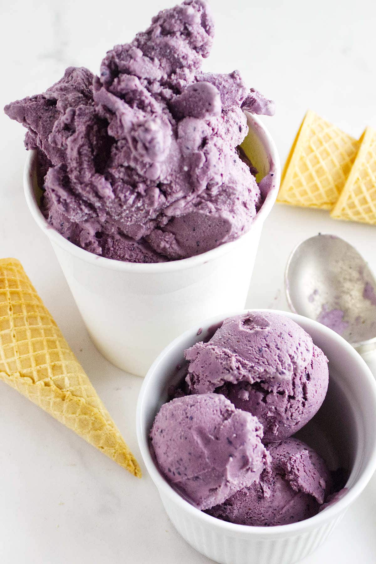 Two containers of ube ice cream-one in a white cup and one in a ramekin-are surrounded by empty waffle cones and a metal ice cream scoop on a white surface, perfect alongside acai berry ice cream for a vibrant dessert duo.
