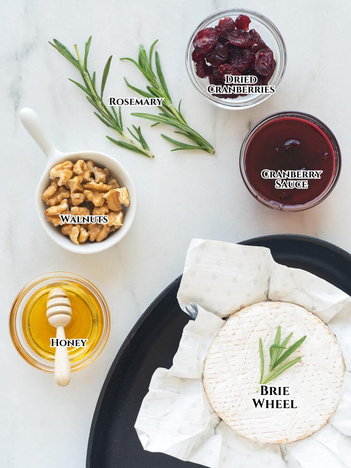 Ingredients for baked Brie arranged on a white surface: a sprig of rosemary, a small bowl of dried cranberries, a bowl of cranberry sauce, walnuts, honey with a dipper, and a Brie wheel on parchment paper.