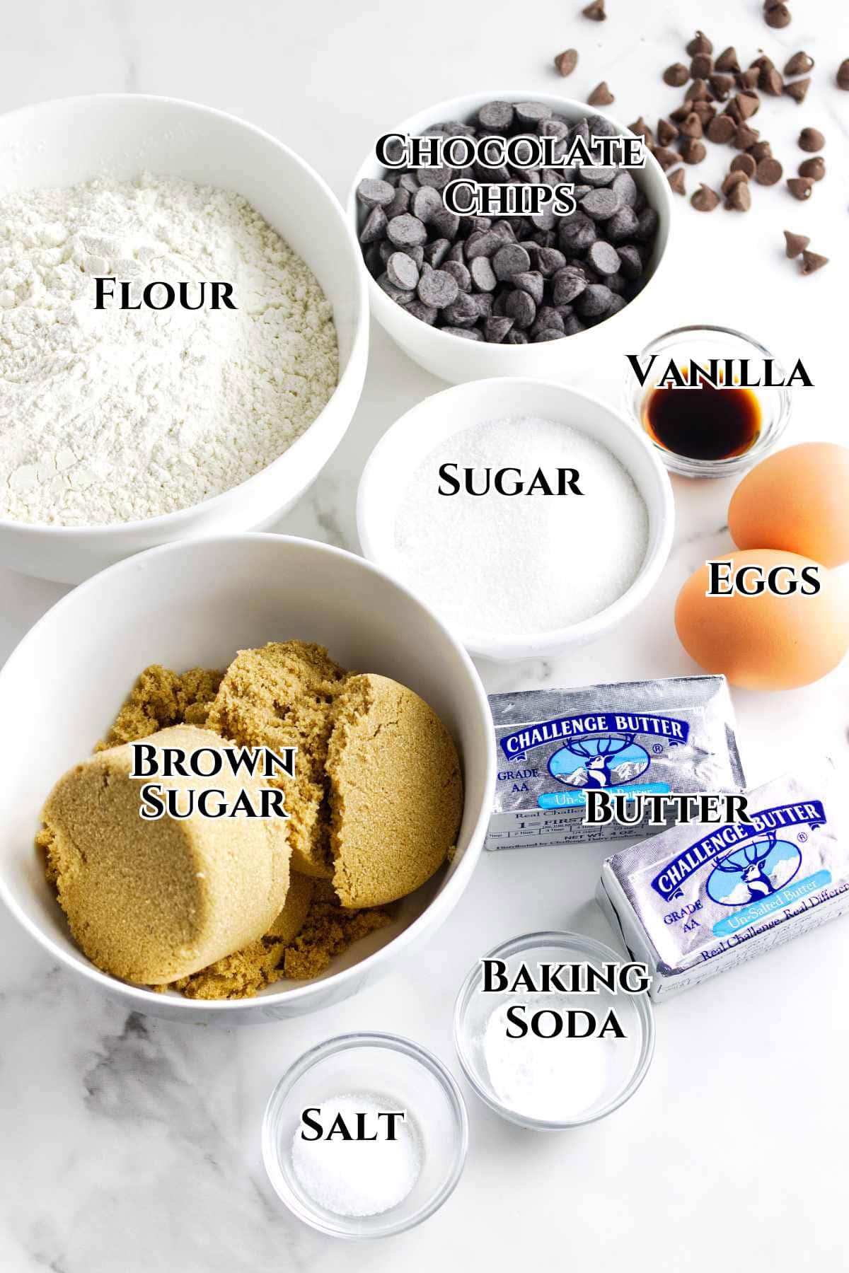 Various chocolate chip cookie ingredients arranged on a white surface: white flour, chocolate chips, granulated sugar, brown sugar, butter sticks, eggs, vanilla extract, baking soda, and salt, each labeled with text.