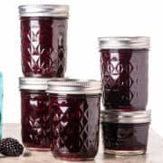 Five glass jars filled with rich, dark purple blackberry preserves are arranged on a countertop. The jars feature silver lids and a diamond pattern on the glass, set against a plain white background.