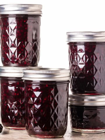Five glass jars filled with rich, dark purple blackberry preserves are arranged on a countertop. The jars feature silver lids and a diamond pattern on the glass, set against a plain white background.