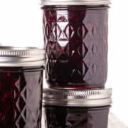 Three glass jars filled with rich blackberry preserves are stacked against a white background. The jars feature silver metal lids and a diamond pattern etched into the glass.