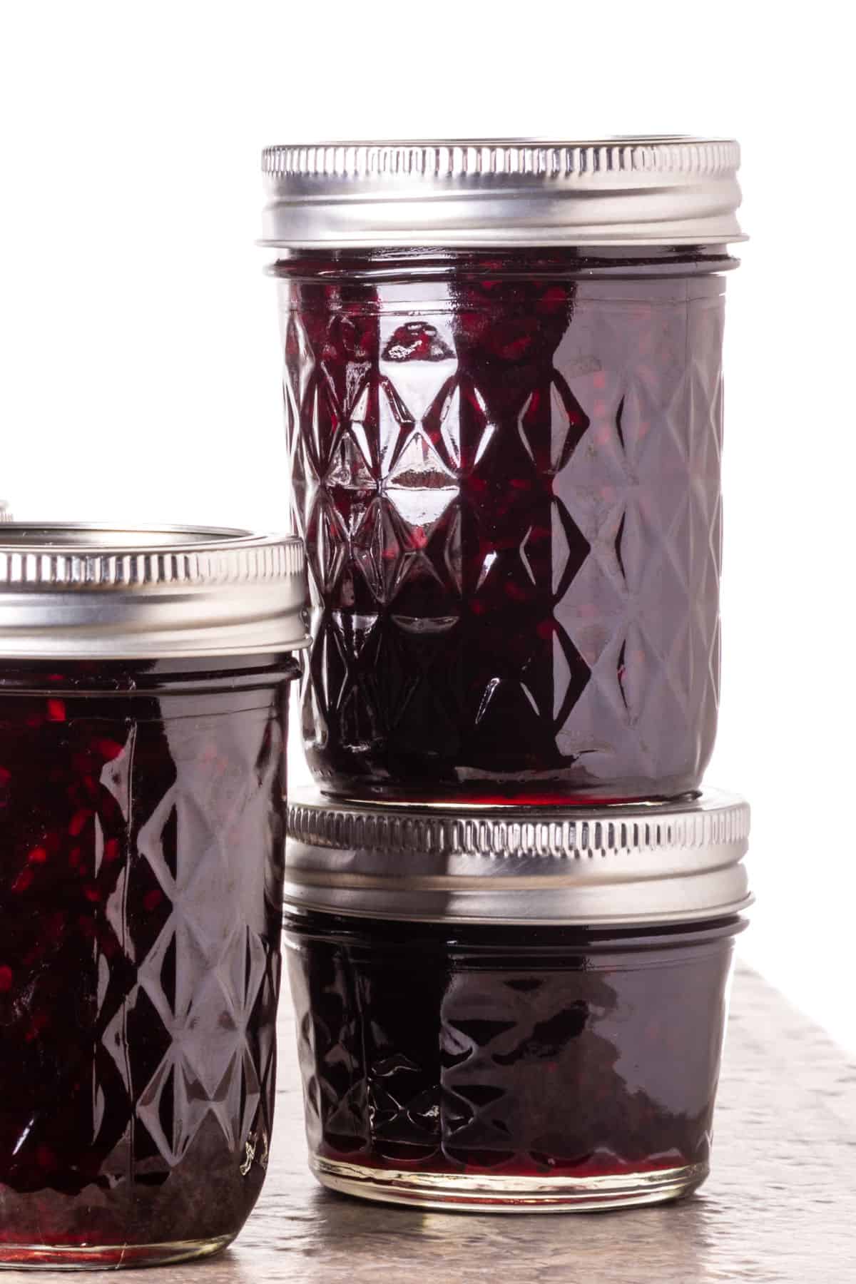 Three glass jars filled with rich blackberry preserves are stacked against a white background. The jars feature silver metal lids and a diamond pattern etched into the glass.