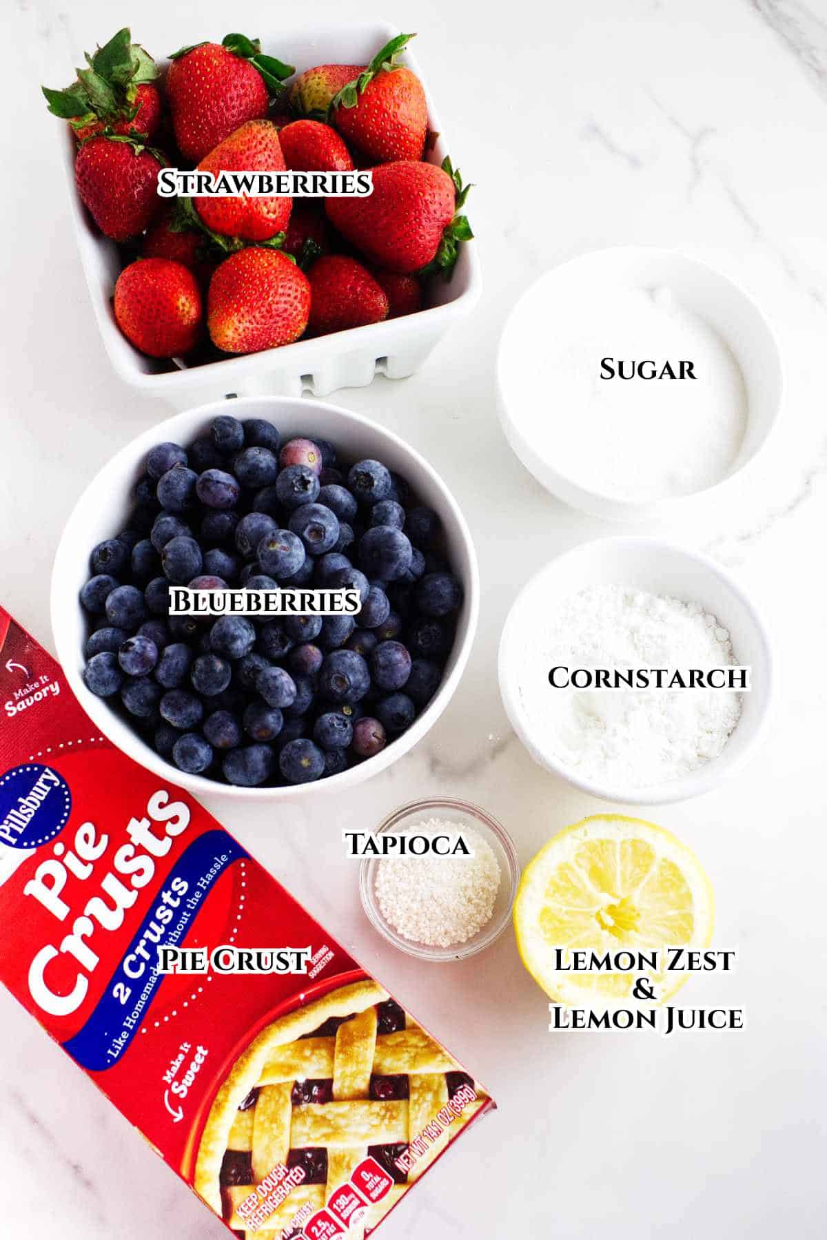 Overhead view of ingredients for a berry pie: fresh strawberries, blueberries, sugar, cornstarch, tapioca, lemon zest and juice, and a package of refrigerated pie crusts on a white surface. Each item is labeled.