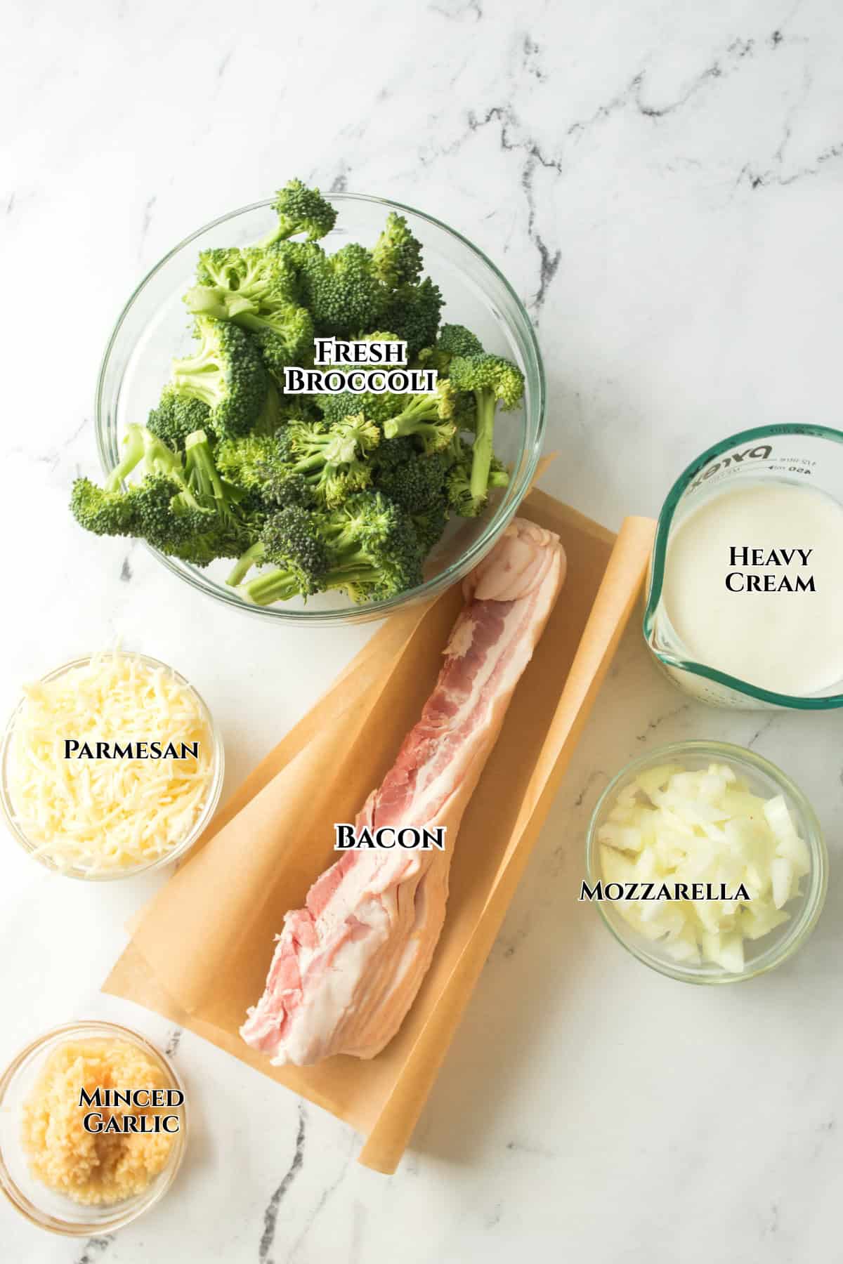A flat lay of broccoli alfredo ingredients on a white surface: fresh broccoli, heavy cream, chopped mozzarella, onions, minced garlic, shredded Parmesan cheese, and a slab of raw bacon on parchment paper.
