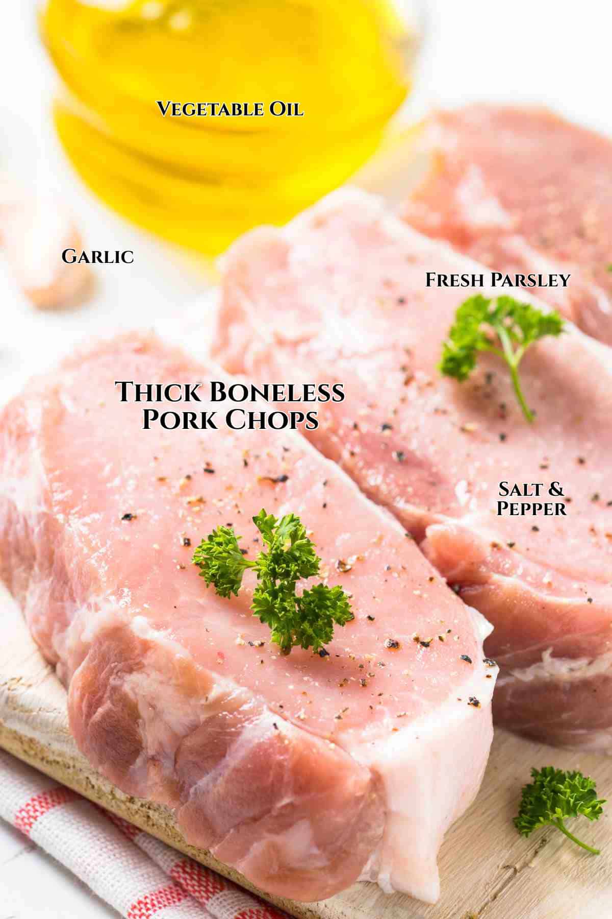 Two thick, boneless pork chops on a cutting board topped with fresh parsley, sprinkled with salt and pepper. Ingredients for making cast iron seared pork chops—garlic cloves and vegetable oil—are labeled nearby.