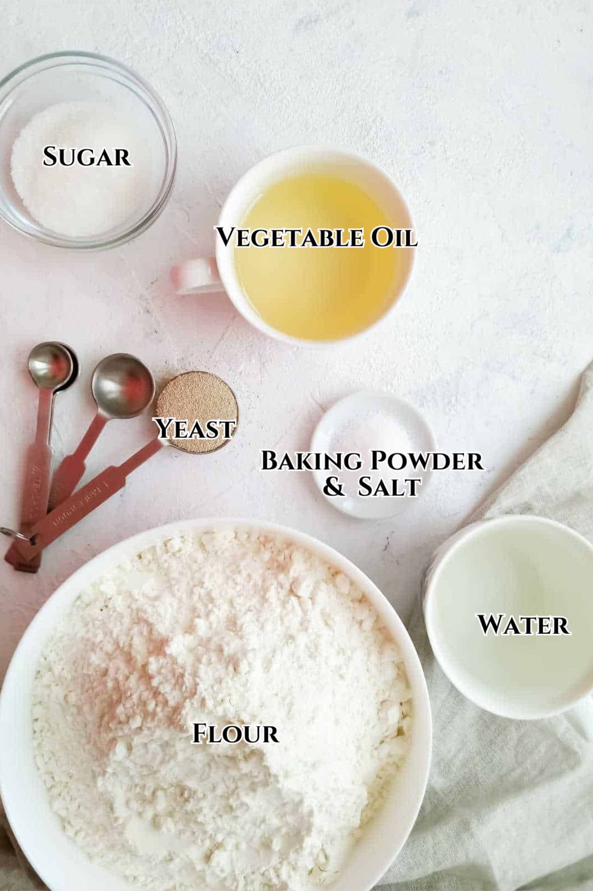 A flat lay of labeled baking ingredients: a bowl of flour, a cup of vegetable oil, a small bowl of sugar, a bowl of water, a dish with baking powder and salt, measuring spoons, and a small dish of yeast on a light surface.