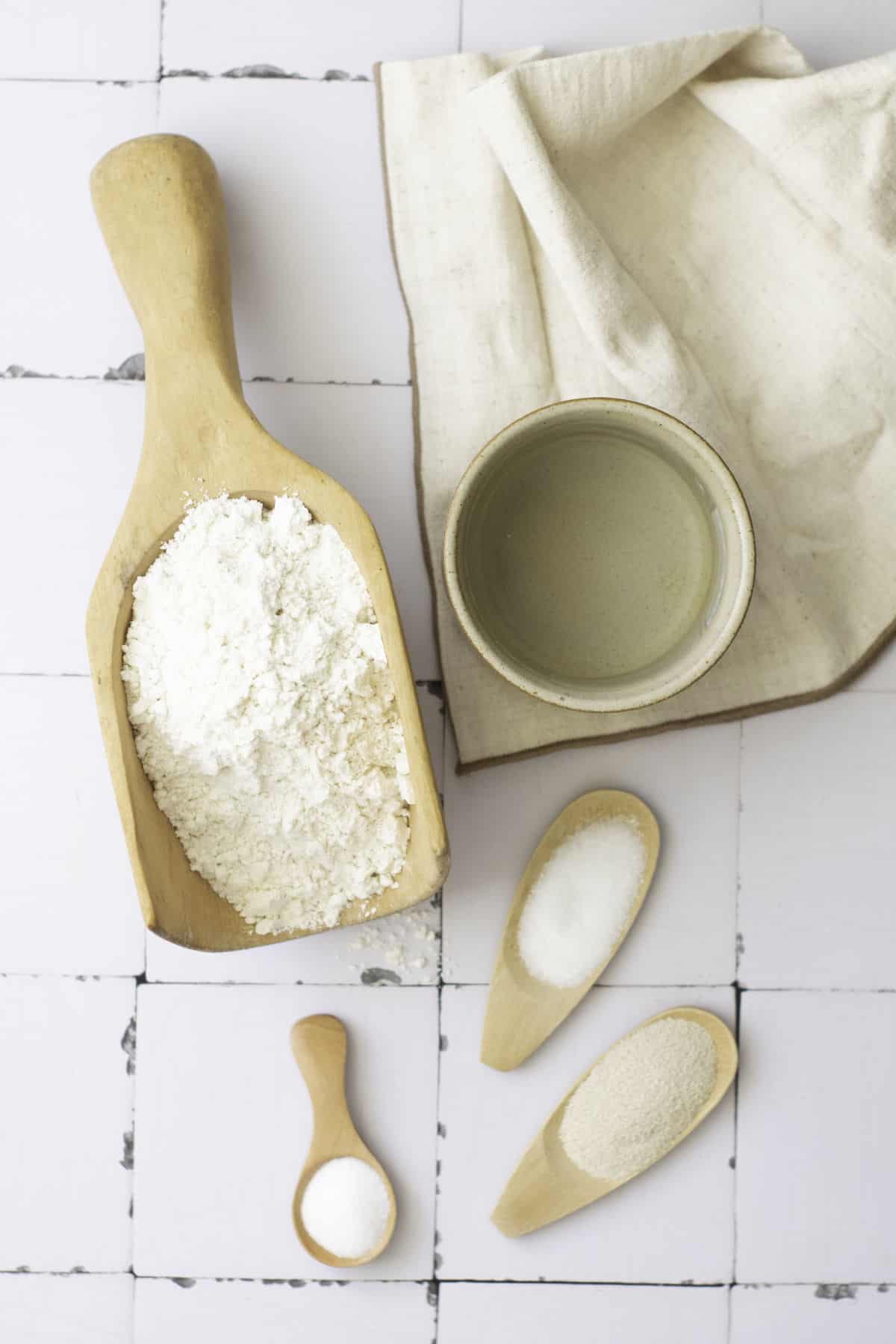 A wooden scoop of flour, a small ceramic bowl with liquid, a beige cloth, and three wooden spoons holding salt, sugar, and yeast are arranged on a white tiled surface—perfect essentials for an easy baguette recipe.