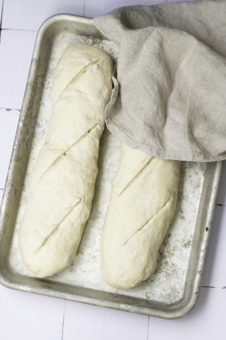Two unbaked baguette loaves with diagonal slashes rest on a metal baking sheet, ready for an easy baguette recipe. A beige cloth partially covers the top right corner of the tray, set on a white surface.