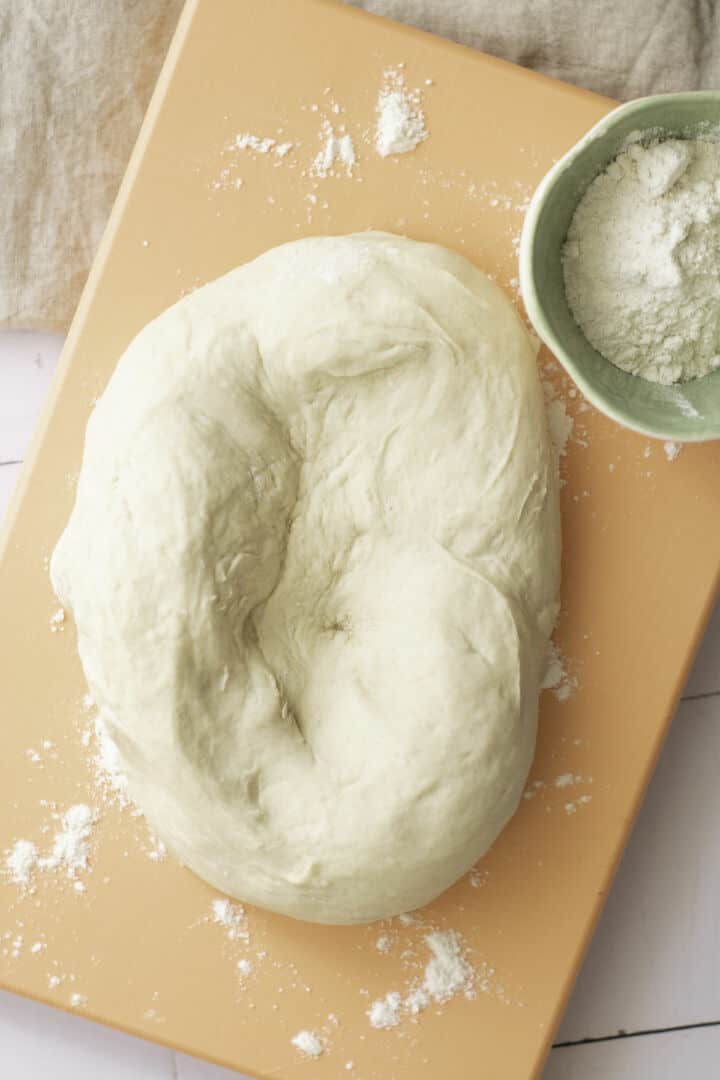 A ball of bread dough rests on a peach-colored cutting board, with scattered flour nearby-a perfect start for any easy baguette recipe, complemented by a small green bowl filled with flour.