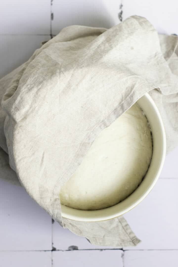 A bowl of dough rising under a light beige cloth on a white surface, partially covered—just the first step in an easy baguette recipe.