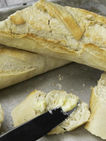 A crusty baguette made from an easy baguette recipe rests on a baking tray, with a few slices cut off. A knife is spreading butter onto one of the slices.