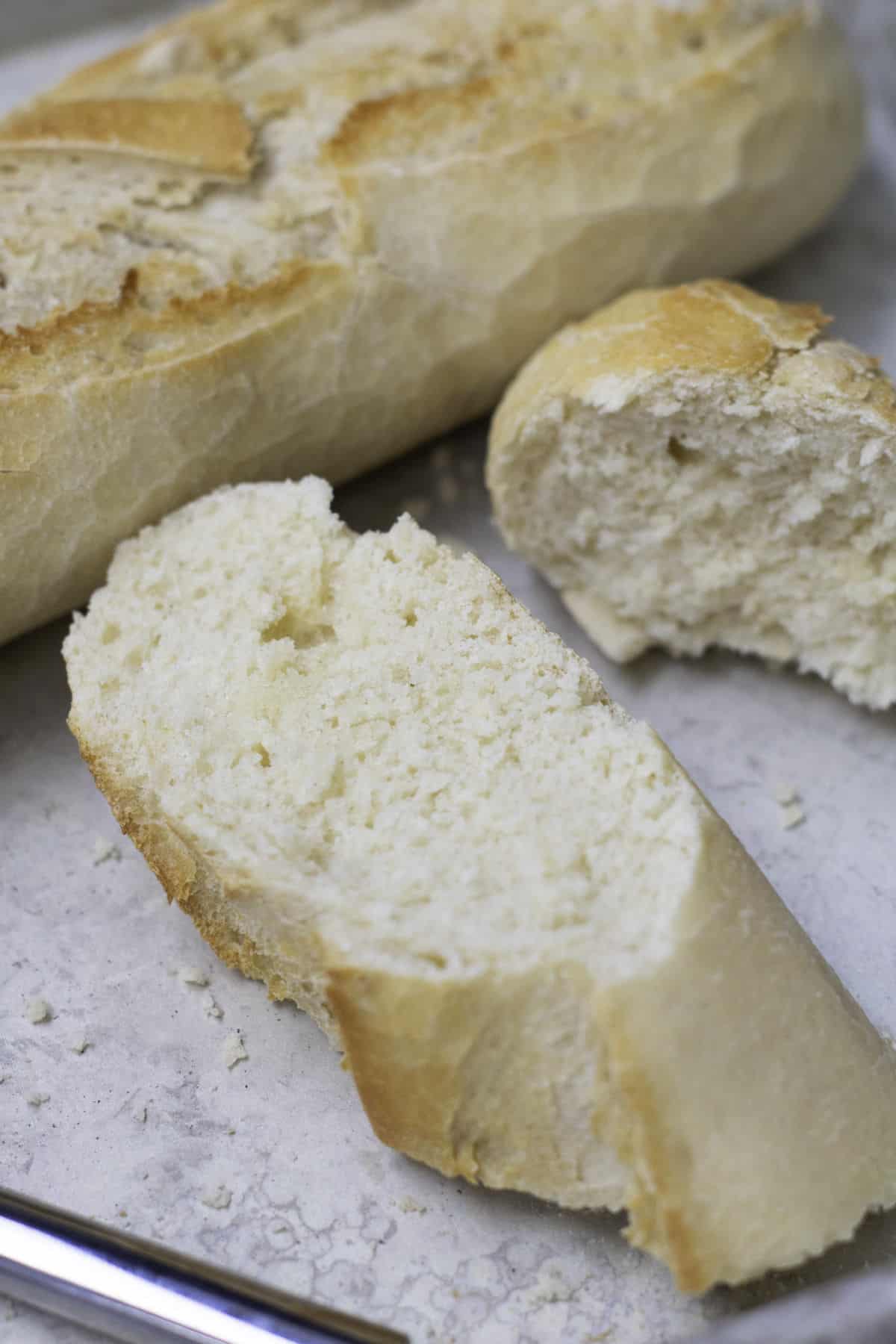 A close-up of a sliced baguette made from an easy baguette recipe on a light-colored surface, showing its golden-brown crust and soft, airy interior. Crumbs are scattered around the bread.