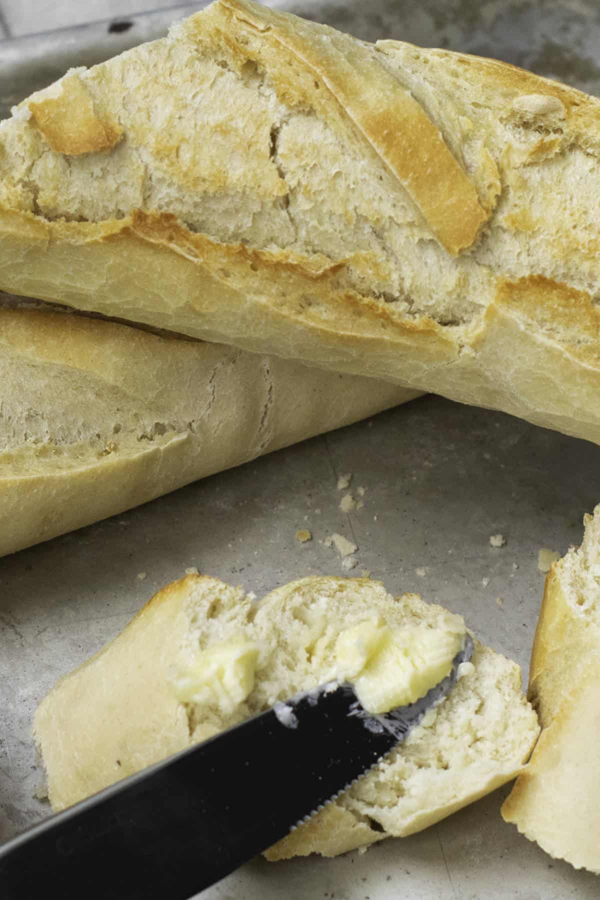 A close-up of a sliced baguette on a metal tray, with a knife spreading butter onto one of the pieces—perfect for serving after trying an easy baguette recipe.