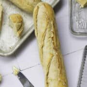 A freshly baked baguette on a white tiled surface, made from an easy baguette recipe, with a baking tray holding another baguette and a bread piece. Nearby are butter pats in a glass dish and a butter knife with a dab of butter.
